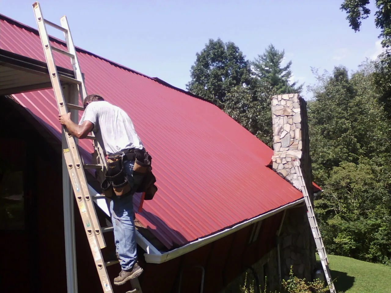 Roofer on a ladder beside a red metal roof and stone chimney — Soffit & Fascia Repair by Roofing Brothers in Talbotton